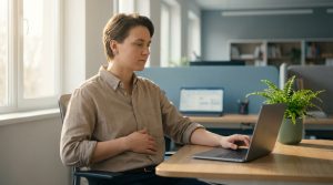 Femme au bureau, main sur la poitrine, travaillant sur un ordinateur portable. Exprime un possible inconfort post-fracture du sternum.