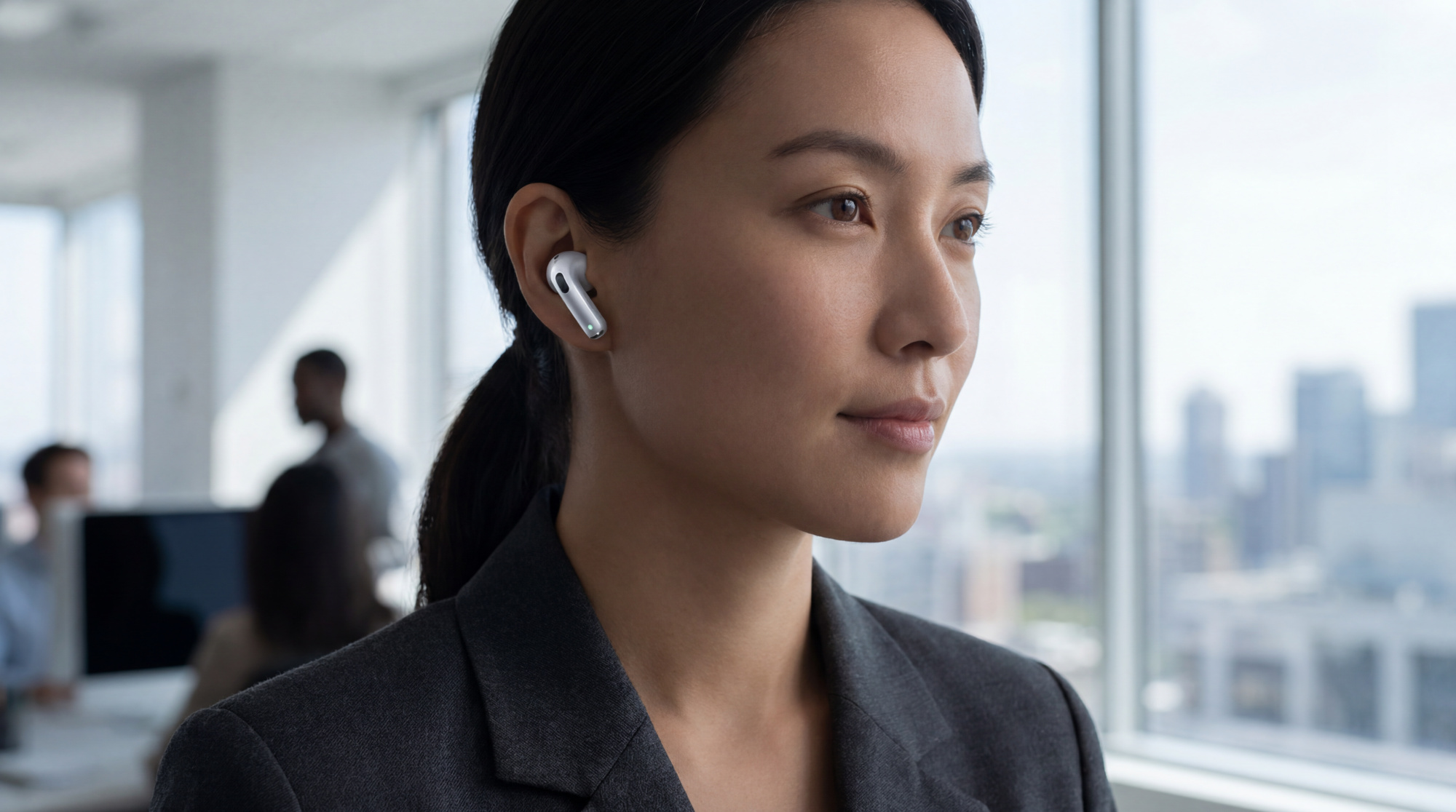 Femme asiatique portant un écouteur sans fil Apple (AirPod) dans un bureau moderne avec vue sur la ville. Des collègues sont flous derrière.